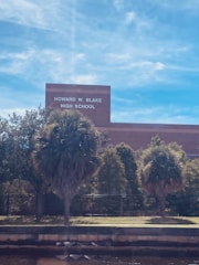 A building with the sign 'Howard W. Blake High School' is visible. The foreground has several palm trees and shrubs, with a fence running parallel to the building. The sky is clear with a few wispy clouds.