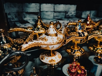 A collection of intricately designed ornate lamps with golden metallic patterns and embellishments sits on a reflective surface. The lamps are adorned in various colors including white, navy blue, and red, all featuring elaborate gold detailing and reflective surfaces. The background is slightly blurred, focusing attention on the detailed craftsmanship of the lamps.