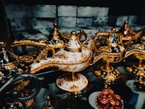 A collection of intricately designed ornate lamps with golden metallic patterns and embellishments sits on a reflective surface. The lamps are adorned in various colors including white, navy blue, and red, all featuring elaborate gold detailing and reflective surfaces. The background is slightly blurred, focusing attention on the detailed craftsmanship of the lamps.