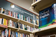 A bookshelf filled with a variety of books, primarily in English. The visible sign above the shelf indicates these are books in English, and several shelves are labeled with alphabetical markers. A large dictionary is prominently displayed, suggesting a focus on English language resources.