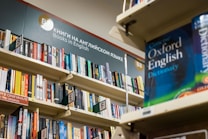 A bookshelf filled with a variety of books, primarily in English. The visible sign above the shelf indicates these are books in English, and several shelves are labeled with alphabetical markers. A large dictionary is prominently displayed, suggesting a focus on English language resources.