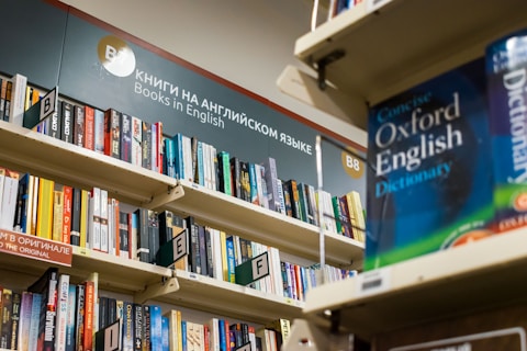 A bookshelf filled with a variety of books, primarily in English. The visible sign above the shelf indicates these are books in English, and several shelves are labeled with alphabetical markers. A large dictionary is prominently displayed, suggesting a focus on English language resources.