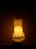 A glowing, sculpted object resembling a sitting animal, likely a dog or a cat, is illuminated from within. The light is warm and softly diffused, casting a gentle glow against a dark background.