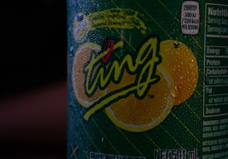 A close-up view of a beverage can covered in condensation droplets. The label features the word 'Ting' prominently in stylized, playful script with vibrant lemon graphics in the background. Nutritional information is partially visible on the side of the can.