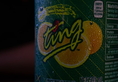 A close-up view of a beverage can covered in condensation droplets. The label features the word 'Ting' prominently in stylized, playful script with vibrant lemon graphics in the background. Nutritional information is partially visible on the side of the can.