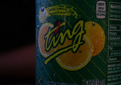 A close-up view of a beverage can covered in condensation droplets. The label features the word 'Ting' prominently in stylized, playful script with vibrant lemon graphics in the background. Nutritional information is partially visible on the side of the can.