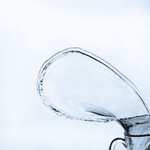 A large, clear splash of water arcs gracefully from a glass container, capturing motion at its peak. The fluidity of the water and the transparency of the container create a dynamic and elegant visual effect against a soft, light background.