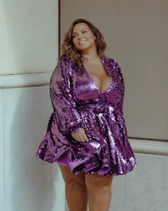 A stylish woman wearing a flowing purple evening gown with gold accents, standing confidently in a softly lit boutique.