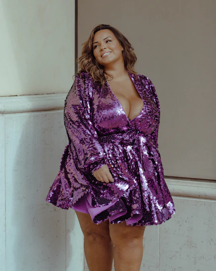 A stylish woman wearing a flowing purple evening gown with gold accents, standing confidently in a softly lit boutique.