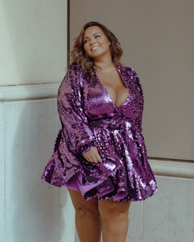 A person stands confidently in a sequined purple dress with long curly hair, leaning slightly against a light-colored wall. The dress has a deep V-neck design and reflects light, adding a vibrant touch to the scene. The person appears to be smiling warmly, creating a friendly and approachable atmosphere.