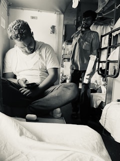 A man sits cross-legged on a train berth, focused on his mobile phone. Another man stands nearby in a corridor, holding a rolled-up item, possibly a newspaper or bedroll. The setting is inside a train compartment with visible seat numbers and bedding.
