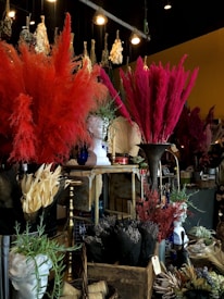 A richly decorated interior with large arrangements of colorful dried flowers, including vibrant reds and pinks. A classical-style white bust is prominently displayed among the florals. Various ornamental items, such as vases and baskets, are scattered throughout, creating a dense and textured visual experience. The lighting provides a warm, atmospheric glow, highlighting the intricate details.