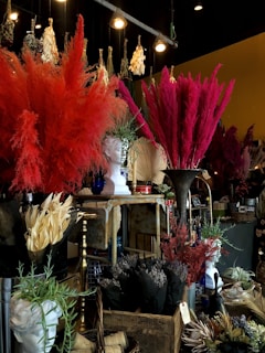 A richly decorated interior with large arrangements of colorful dried flowers, including vibrant reds and pinks. A classical-style white bust is prominently displayed among the florals. Various ornamental items, such as vases and baskets, are scattered throughout, creating a dense and textured visual experience. The lighting provides a warm, atmospheric glow, highlighting the intricate details.