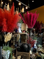 A richly decorated interior with large arrangements of colorful dried flowers, including vibrant reds and pinks. A classical-style white bust is prominently displayed among the florals. Various ornamental items, such as vases and baskets, are scattered throughout, creating a dense and textured visual experience. The lighting provides a warm, atmospheric glow, highlighting the intricate details.