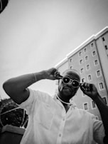 A person wearing dark sunglasses and a light-colored shirt is posing confidently outdoors. The background features a tall building with multiple windows and a clear sky. The photographic style is black and white, contributing to an urban and classic feel.