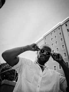 A person wearing dark sunglasses and a light-colored shirt is posing confidently outdoors. The background features a tall building with multiple windows and a clear sky. The photographic style is black and white, contributing to an urban and classic feel.