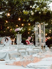 a table with white tables and chairs under a tree with lights