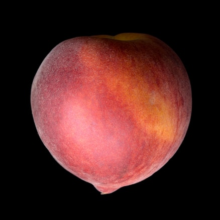 A ripe peach with a smooth, velvety surface is centered against a stark black background. The peach displays varying shades of red, yellow, and orange, highlighting its freshness and natural texture.
