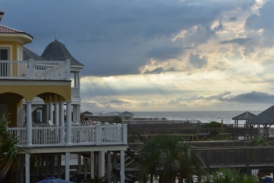 A seaside scene with a multi-story house featuring a yellow and white exterior. The house has multiple balconies overlooking a wooden boardwalk and palm trees. In the distance, there is a cloudy sky with beams of sunlight breaking through, over an expansive view of the ocean.