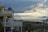 A seaside scene with a multi-story house featuring a yellow and white exterior. The house has multiple balconies overlooking a wooden boardwalk and palm trees. In the distance, there is a cloudy sky with beams of sunlight breaking through, over an expansive view of the ocean.