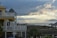 A seaside scene with a multi-story house featuring a yellow and white exterior. The house has multiple balconies overlooking a wooden boardwalk and palm trees. In the distance, there is a cloudy sky with beams of sunlight breaking through, over an expansive view of the ocean.