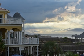 A seaside scene with a multi-story house featuring a yellow and white exterior. The house has multiple balconies overlooking a wooden boardwalk and palm trees. In the distance, there is a cloudy sky with beams of sunlight breaking through, over an expansive view of the ocean.
