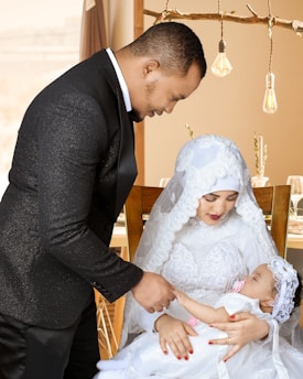 A man dressed in a black suit leans gently toward a seated woman in a white lace dress, holding a sleeping baby also dressed in white. The setting has a warm ambiance with decorative hanging lights and a softly blurred background.