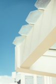 A modern architectural feature with geometric, hexagonal glass panels framed against a clear blue sky. The structure is composed of light-colored materials, creating a minimalist aesthetic.