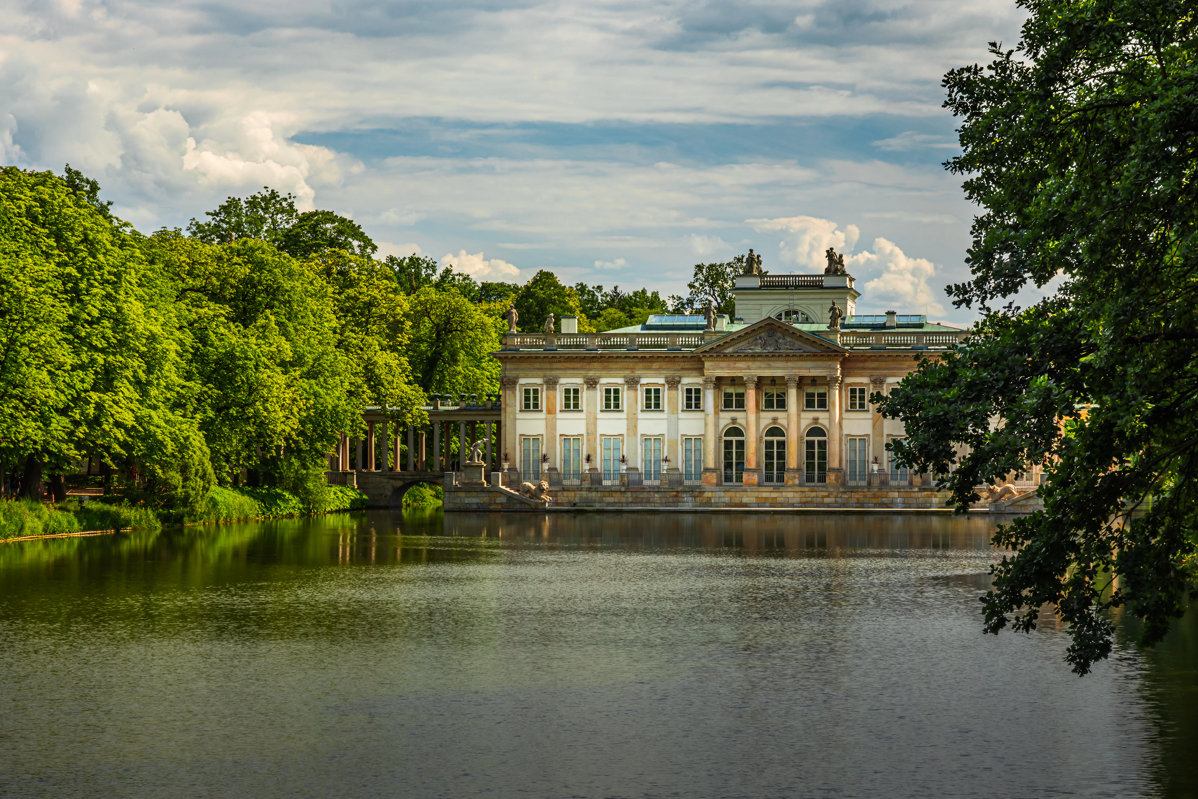 The neoclassical Palace on the Isle in Warsaw's Lazienki Park seen across the still lake in summer, its columned front framed by heavy green trees under a sky of tall white cloud