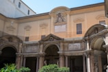 An architectural facade featuring classical design elements, including arched openings, columns, and decorative reliefs. The structure is adorned with a central plaque and crest, and the walls are painted in a warm beige tone. Lush greenery is visible in the foreground, adding a touch of nature to the scene.