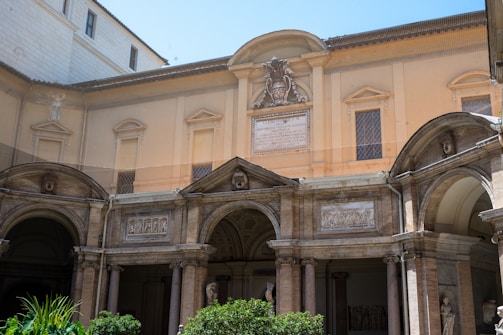 An architectural facade featuring classical design elements, including arched openings, columns, and decorative reliefs. The structure is adorned with a central plaque and crest, and the walls are painted in a warm beige tone. Lush greenery is visible in the foreground, adding a touch of nature to the scene.