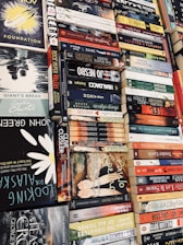 A collection of various books piled horizontally and vertically, showcasing titles from a range of genres. There are both classic and contemporary book covers with vibrant colors and diverse typographic styles.