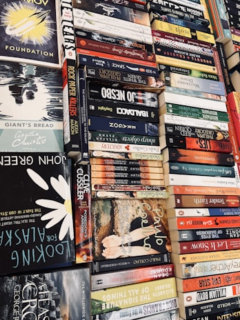 A collection of various books piled horizontally and vertically, showcasing titles from a range of genres. There are both classic and contemporary book covers with vibrant colors and diverse typographic styles.