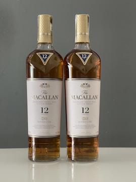 Two bottles of Macallan Highland Single Malt Scotch Whisky, 12 Years Old Double Cask, are placed side by side against a plain background. Both bottles are sealed and have a consistent label design featuring gold and white colors.