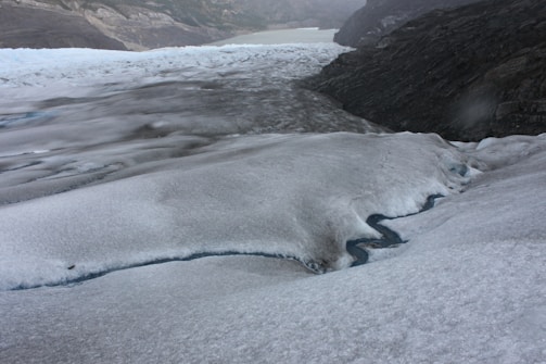 A vast, icy landscape featuring an expansive glacier with visible crevasses and a small stream of melted water flowing through. In the background, rugged mountains and a hazy sky create a dramatic, remote atmosphere.