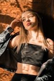 A woman with long blonde hair stands against a wooden background. Sunlight filters through leaves, casting dappled shadows over her face and body. She wears a black top and a denim jacket that has fallen off her shoulders. Her expression is contemplative, looking slightly upward and to the side.