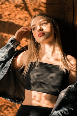 A woman with long blonde hair stands against a wooden background. Sunlight filters through leaves, casting dappled shadows over her face and body. She wears a black top and a denim jacket that has fallen off her shoulders. Her expression is contemplative, looking slightly upward and to the side.