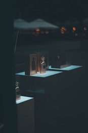Several perfume bottles and their packaging are displayed on illuminated stands against a dark background. Subtle blurred lights in the background create a soft bokeh effect, adding a sophisticated atmosphere.