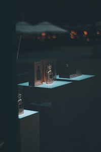 Several perfume bottles and their packaging are displayed on illuminated stands against a dark background. Subtle blurred lights in the background create a soft bokeh effect, adding a sophisticated atmosphere.