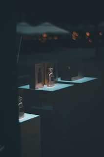 Several perfume bottles and their packaging are displayed on illuminated stands against a dark background. Subtle blurred lights in the background create a soft bokeh effect, adding a sophisticated atmosphere.