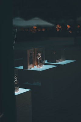 Several perfume bottles and their packaging are displayed on illuminated stands against a dark background. Subtle blurred lights in the background create a soft bokeh effect, adding a sophisticated atmosphere.