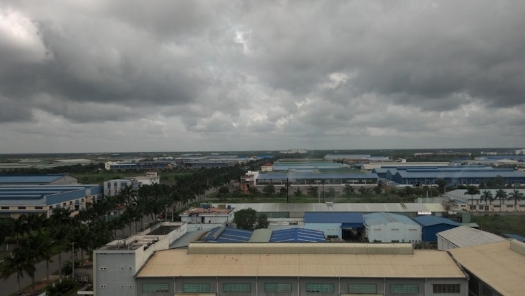 A vast industrial area with large blue-roofed buildings surrounded by greenery and palm trees under a cloudy sky. The landscape stretches far with factories and warehouses dominating the scene.