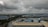 A vast industrial area with large blue-roofed buildings surrounded by greenery and palm trees under a cloudy sky. The landscape stretches far with factories and warehouses dominating the scene.