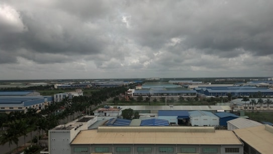 A vast industrial area with large blue-roofed buildings surrounded by greenery and palm trees under a cloudy sky. The landscape stretches far with factories and warehouses dominating the scene.
