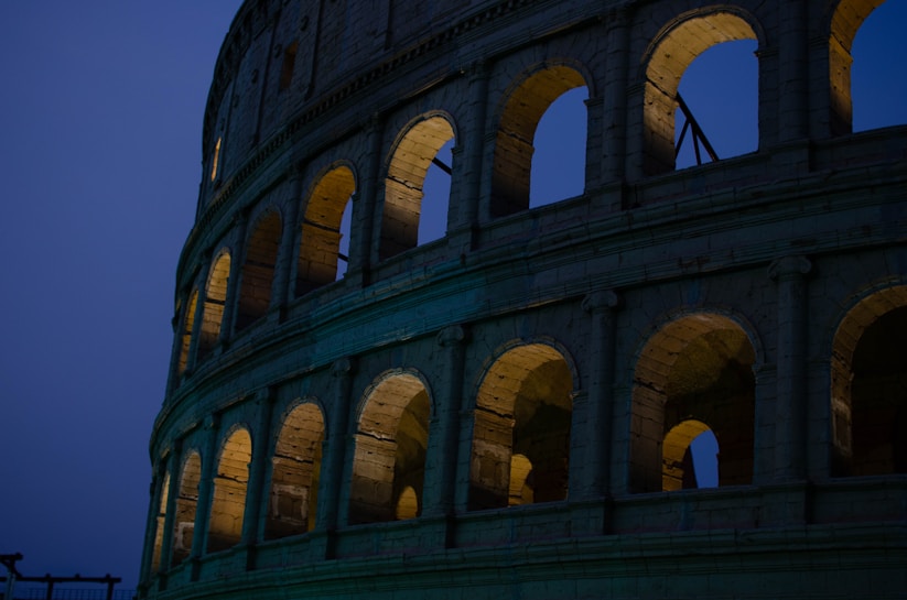 The image captures the ancient Colosseum in Rome during twilight, partially illuminated from within. The curved structure showcases rows of arches, revealing the monumental architectural design against a deep blue sky.