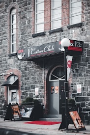 A brick building with a sign for the 'Cotton Club', an adult entertainment venue. The entrance features a red carpet leading to the door, with promotional posters displayed on stands outside. A streetlamp with a round light is positioned near the entrance.