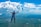 A skydiver in a blue jumpsuit and helmet is free-falling above a vast landscape of green fields. Another skydiver is visible in the distance. Large, fluffy clouds dominate the bright blue sky, casting shadows on the ground below.