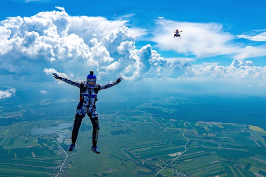 A skydiver in a blue jumpsuit and helmet is free-falling above a vast landscape of green fields. Another skydiver is visible in the distance. Large, fluffy clouds dominate the bright blue sky, casting shadows on the ground below.