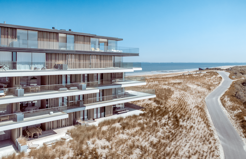 A modern multi-story building with sleek glass balconies overlooks a sandy beach with scattered vegetation. A road winds along the edge of the beach, leading to the horizon. The ocean is calm and the sky is clear and blue.