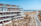A modern multi-story building with sleek glass balconies overlooks a sandy beach with scattered vegetation. A road winds along the edge of the beach, leading to the horizon. The ocean is calm and the sky is clear and blue.